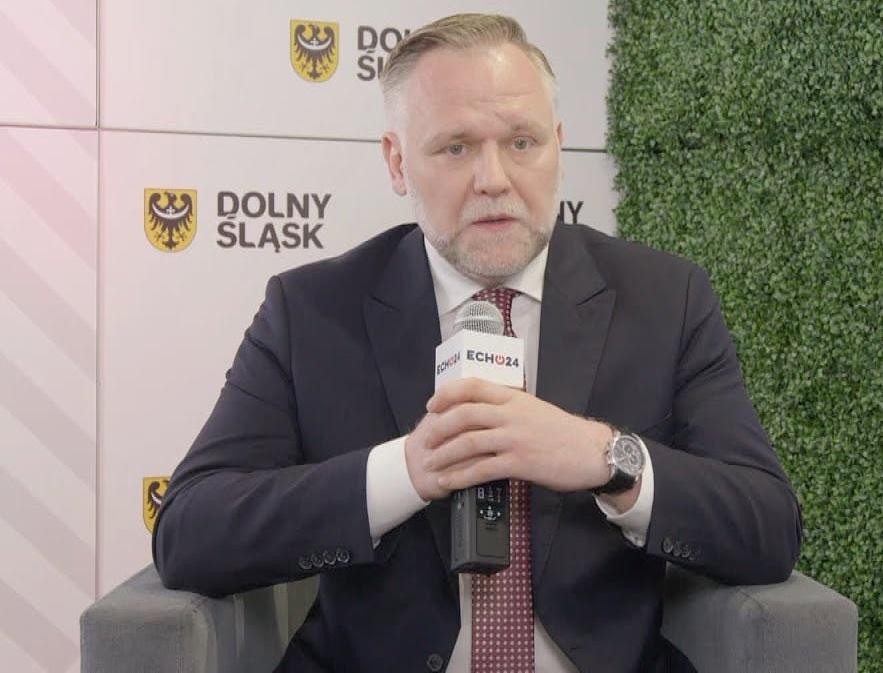 Dawid Jackiewicz przemawiający publicznie, gestykulujący, na tle niewyraźnej publiczności lub sali konferencyjnej.