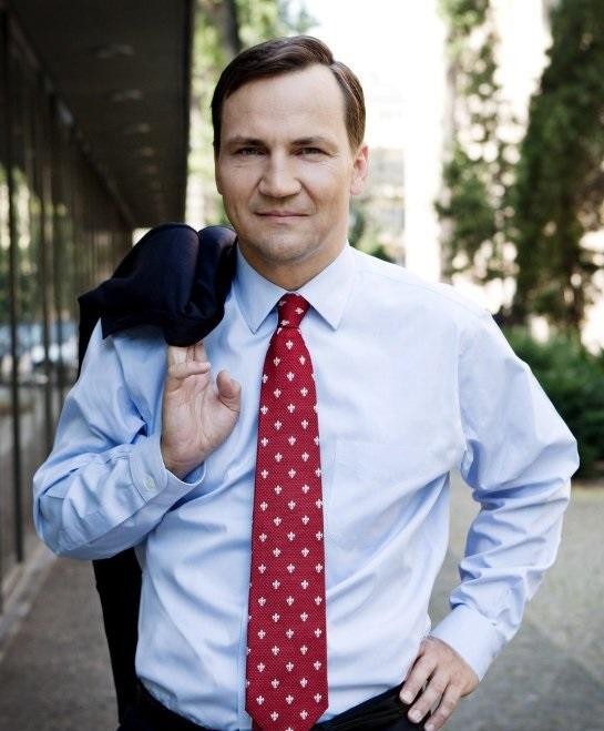 Radosław Sikorski podczas wystąpienia publicznego, gestykulujący, w tle zamazane postaci, oddające dynamikę wydarzenia.